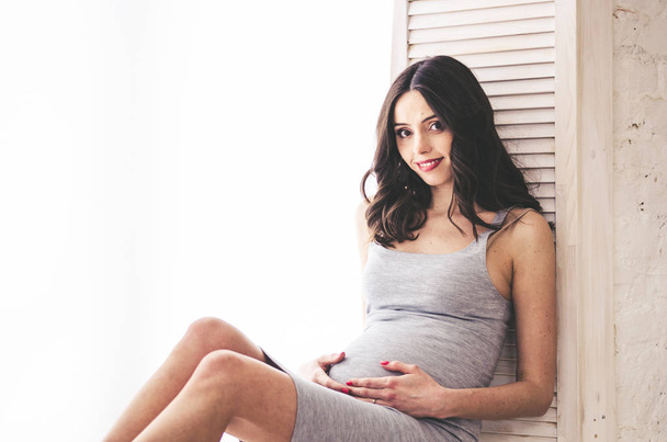 Έγκυος γυναίκα στο γκρι φόρεμα κάθεται κοντά στο παράθυρο - Φωτογραφία, εικόνα