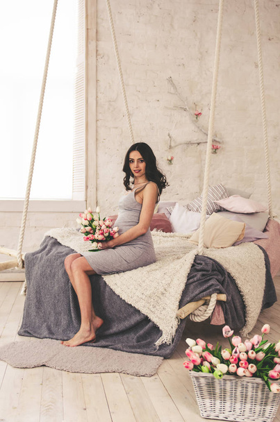 happy pregnant woman in grey dress sitting on bed - Foto, Imagem