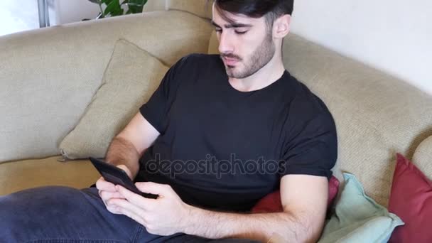 Όμορφος νεαρός άνδρας διαβάζει ebook στον καναπέ - Πλάνα, βίντεο