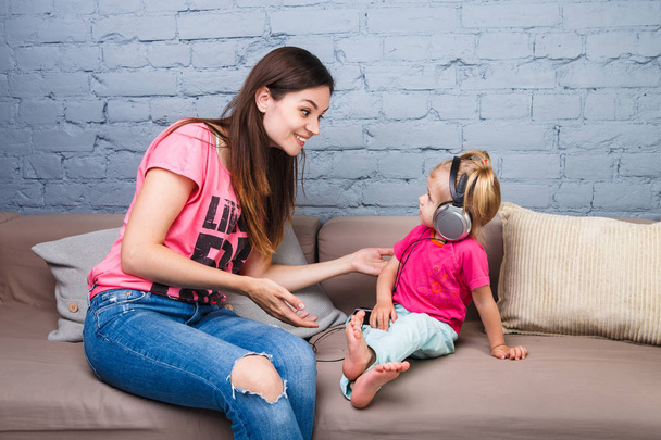 Mamá y su hija escuchan música con grandes auriculares puestos en su cabeza, sentados en el sofá. Sostiene el teléfono
. - Foto, Imagen