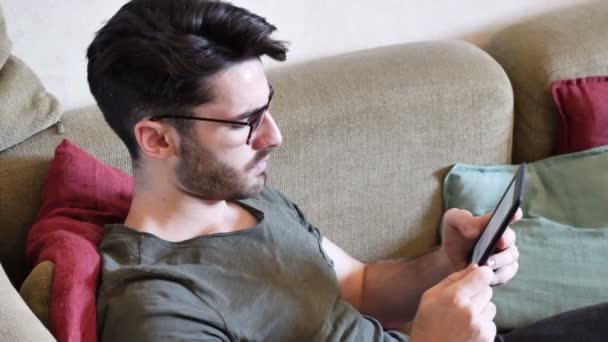 Handsome young man reading ebook on sofa - Felvétel, videó