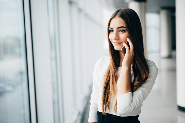 Happy businesswoman standing in her office in a highrise building overlooking the cityscape. Businesswoman talking on mobile phone standing near window in office. - Foto, Imagem