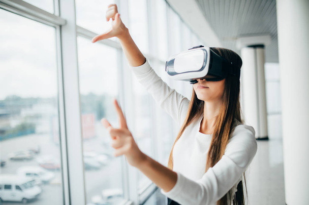 Beautiful girl using virtual reality glasses near bright window with skyscraper view outside. Business woman wearing VR goggles - Photo, Image