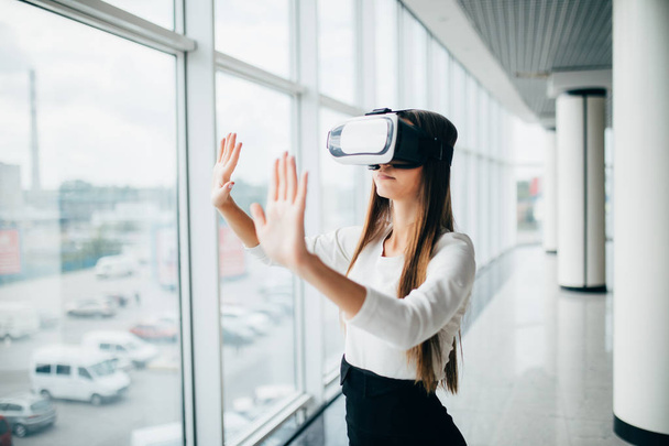 Beautiful girl using virtual reality glasses near bright window with skyscraper view outside. Business woman wearing VR goggles - Photo, Image
