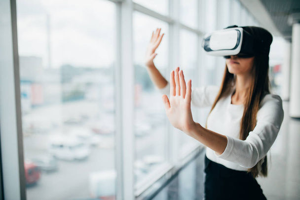 Красива дівчина використовує окуляри віртуальної реальності біля яскравого вікна з видом на хмарочос зовні. Ділова жінка в окулярах VR
 - Фото, зображення
