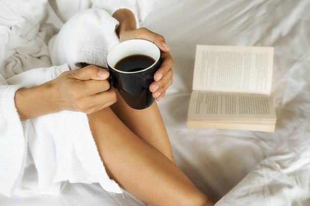 schöne asiatische Frau sitzt auf dem Bett mit einer Tasse Kaffee und liest ein Buch. Morgen mit einem Buch und einer Tasse Kaffee. Entspannungskonzept. Retro-Filtereffekt, weicher Fokus, selektiver Fokus - Foto, Bild