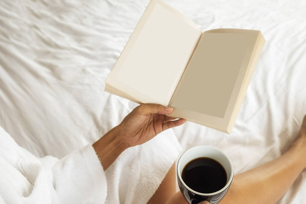 Όμορφη ασιατική γυναίκα κάθεται στο κρεβάτι με ένα φλιτζάνι καφέ και διαβάζοντας ένα βιβλίο. Το πρωί με ένα βιβλίο και το φλιτζάνι του καφέ. Χαλαρωτικό έννοια. Εφέ ρετρό φίλτρου, μαλακή εστίαση, επιλεκτική εστίαση - Φωτογραφία, εικόνα