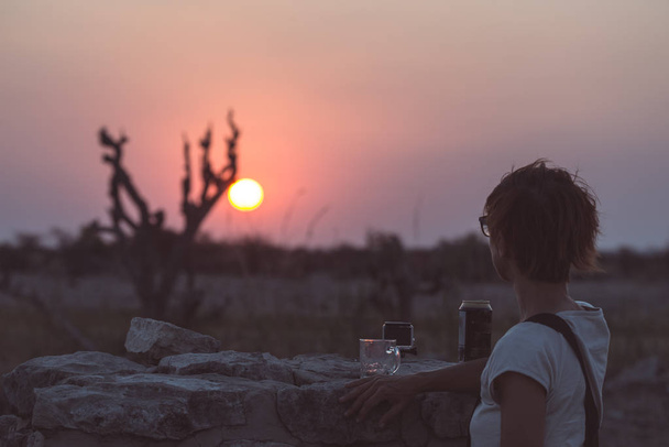 Ένα άτομο με ένα ποτό και να βλέπουν το πολύχρωμο ηλιοβασίλεμα στην έρημο Namib, ταξιδιωτικό προορισμό της Ναμίμπια, στην Αφρική. Έννοια της περιπέτειας και ταξιδεύουν άτομα. - Φωτογραφία, εικόνα