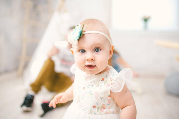 маленька дівчинка один рік з блакитними очима блондинка в пишних біле плаття із задоволенням і грає у світлій кімнаті на фоні Індійська тентова та батьків - Фото, зображення