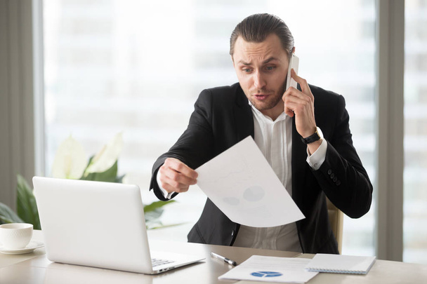 Shocked businessman talks on the phone holding financial report - Fotoğraf, Görsel