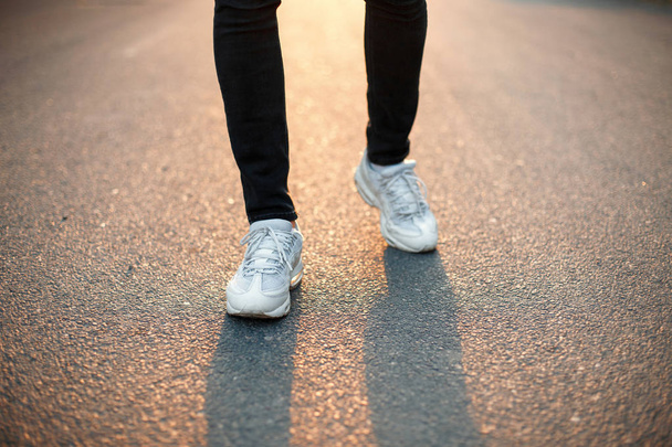 Mužské nohy při chůzi po asfaltové silnici při západu slunce. Bílé tenisky a černé džíny - Fotografie, Obrázek