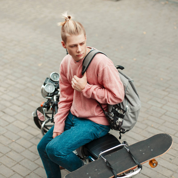 Yakışıklı genç bir sırt çantası ve bir motosiklet üzerinde oturan bir kaykay ile pembe kazaklı bir saç modeli ile - Fotoğraf, Görsel