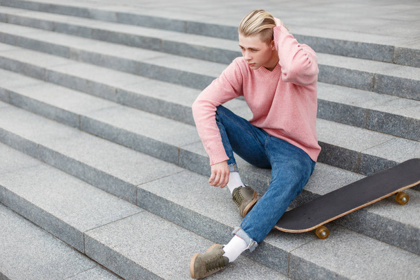 стильный красивый мужчина в модной одежде со скейтбордом сидит на ступеньках
 - Фото, изображение