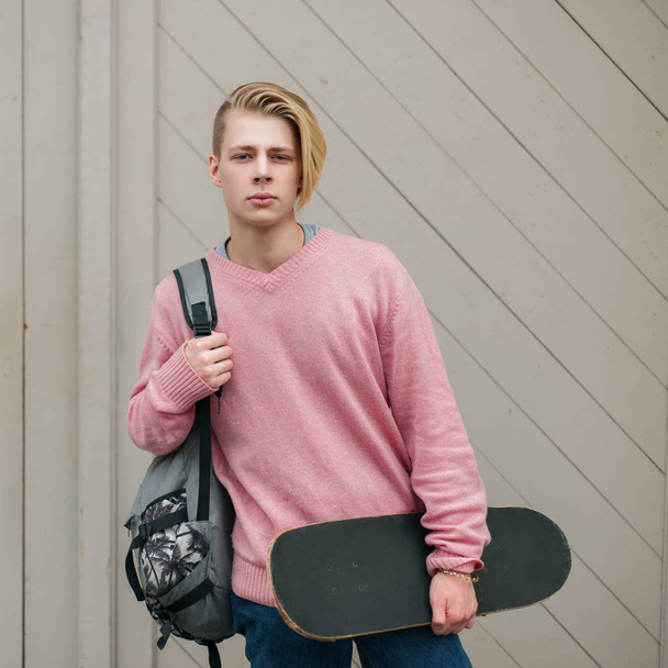Modieuze jonge knappe man in een roze trui met een rugzak en skateboard in de buurt van de houten wand - Foto, afbeelding