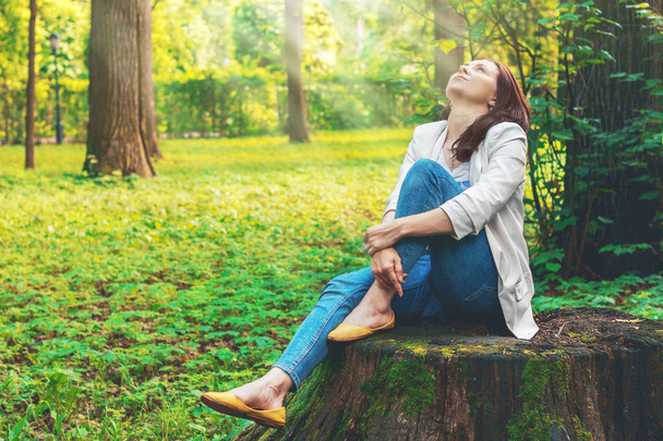 Beautiful girl sits on a big old stump in the forest, the sun's rays breaking through the leaves and illuminate her face. Fabulous country. Cute woman is enjoying of picturesque nature. Camping, rest. - Photo, image