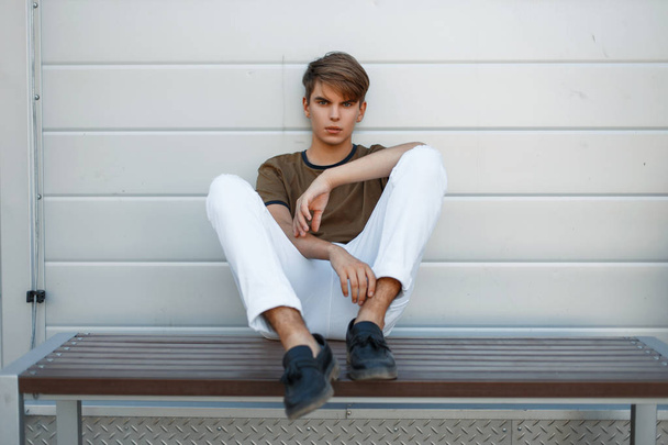 Hombre modelo guapo de moda en una camiseta y pantalones blancos con zapatos negros cerca de la pared de metal brillante
 - Foto, Imagen