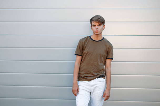 jovem bonito em uma camiseta clássica e calças brancas perto de uma parede de metal cinza
 - Foto, Imagem