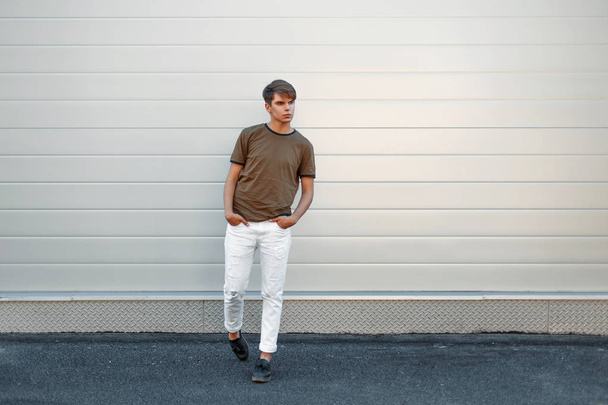 Hermoso joven con estilo en una camiseta de moda y pantalones blancos con pantalones de cuero negro cerca de la pared de metal gris
 - Foto, Imagen