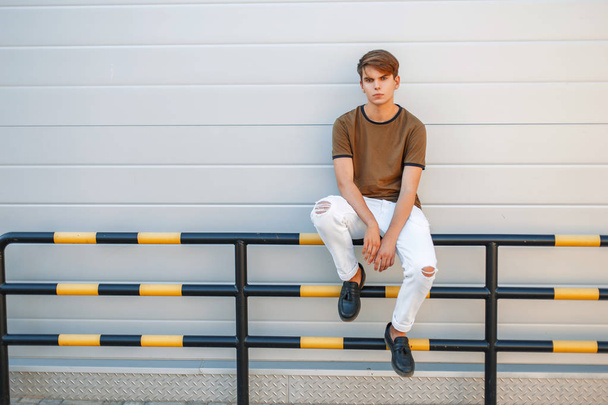 Joven hombre guapo con un peinado en ropa de verano de moda sentado cerca de una pared gris brillante moderno
 - Foto, Imagen