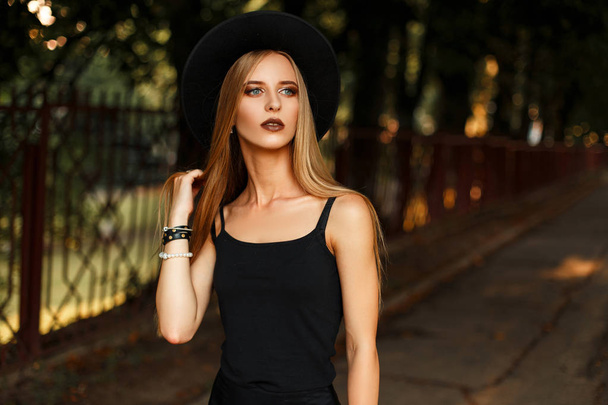 Gyönyörű modell nő elegáns fekete ruhát és divatos fekete kalapban pózol a park - Fotó, kép