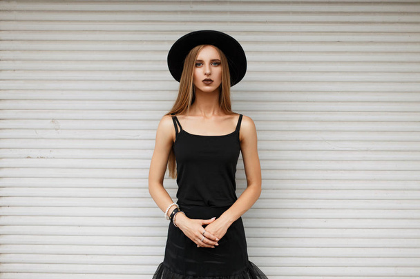 Güzel genç kız trendy siyah şapka ve beyaz bir duvar yakınındaki şık siyah elbise - Fotoğraf, Görsel