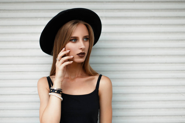 Gyönyörű fiatal modell nő elegáns kalap és divatos ruhák, fehér fém fal közelében - Fotó, kép