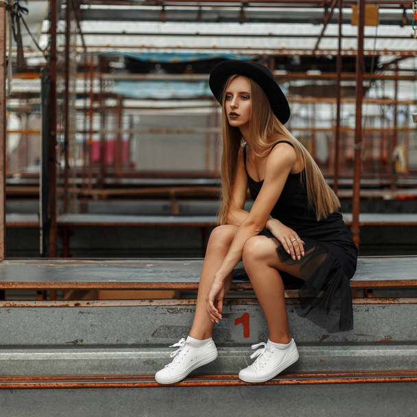 mooie stijlvolle vrouw met een trendy hoed en een zwarte rok met witte schoenen zit op de straat - Foto, afbeelding