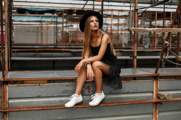 Güzel genç model kadın trendy bir şapka ve beyaz ayakkabı bir masada oturan şık siyah giysili - Fotoğraf, Görsel