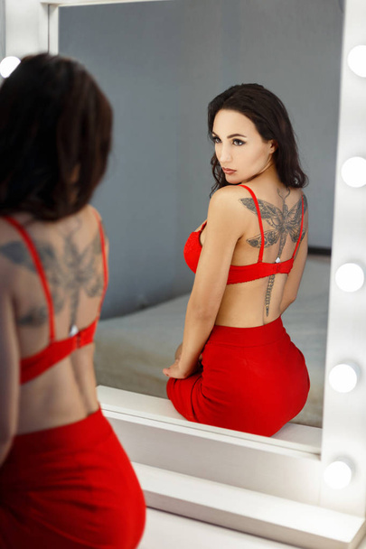 Hermosa joven con un tatuaje en lencería roja sexy se ve en un espejo vintage con bombillas
 - Foto, Imagen