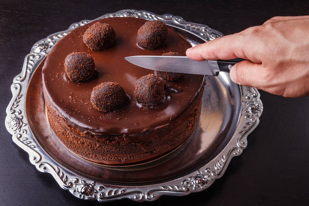 A mans hand with a kitchen knife above a chocolate homemade biscuit cake on a metal plate - Foto, Imagem