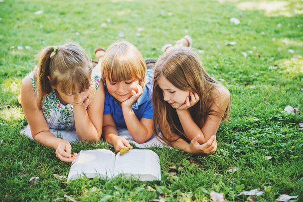 Groupe de trois enfants allongés sur de l'herbe verte et lisant ensemble un livre d'histoire. Éducation des enfants
 - Photo, image