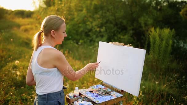 自然の中でキャンバスに彼女の手でブラシを持つ美しい金髪の女性アーティストを描画します - 映像、動画