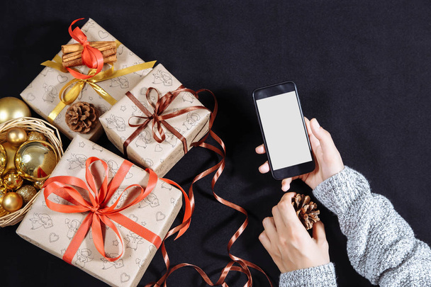 Female hands holding mobile phone on dark background with Christmas gifts. Xmas and Happy New Year composition. Flat lay, top view - Foto, imagen