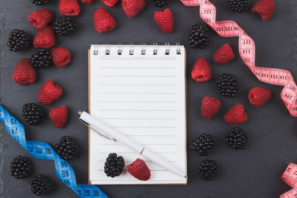 Red raspberries, tape measure, notebook and a pen on the black slate stone background - Photo, Image