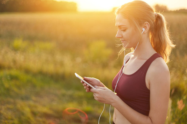 Female runner enjoying calm atmosphere while jogging on field, listening to her favourite track in earphones. Young female going in for sport. People, active lifestyle, health and fitness concept - Foto, immagini