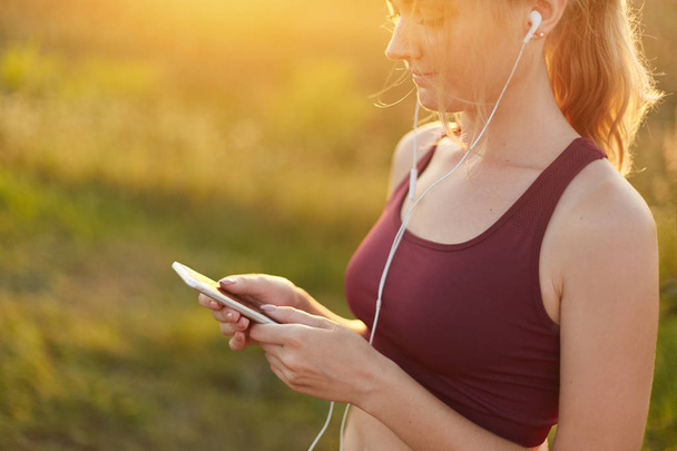 Young sporty woman running, doing sport, wearing top, listening to music with earphones and mobile phone, enjoying fresh air and sunshine. Slim female athlete using modern cell phone. Sport concept - Photo, Image