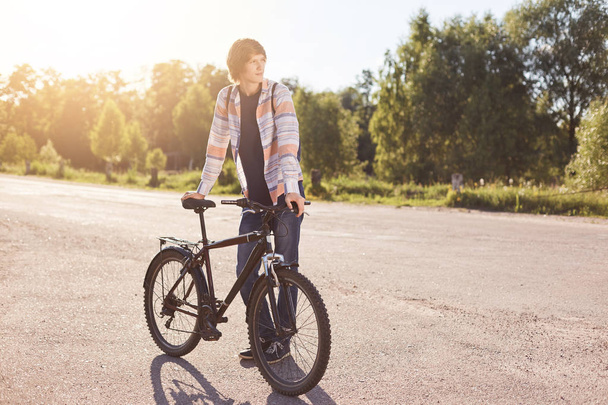 Pensive teenage boy standing on road, keeping hand on handle bar of his bike, waiting for others cyclists to have journey together, looking aside while standing on road. Active lifestyle concept - Photo, Image
