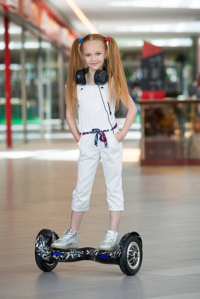Chica feliz y sonriente paseos en mini segway en los auriculares en el centro comercial. Adolescente montando en el hover board o giroscooter y escuchando música
. - Foto, Imagen