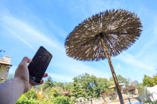 Kädessä pitämällä älypuhelin taustalla tummennettu ranta sateenvarjo
  - Valokuva, kuva
