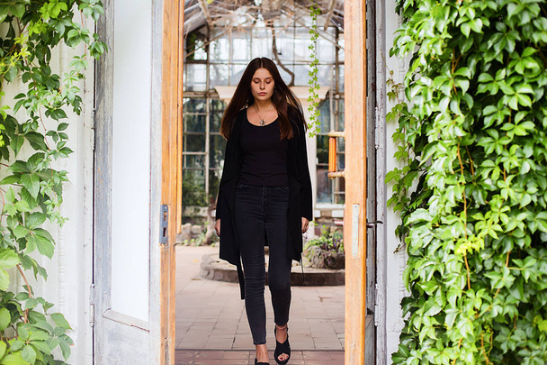 Lifestyle portrait of girl wearing blank black t-shirt, jeans and coat posing against building covered with green leaves - Foto, afbeelding