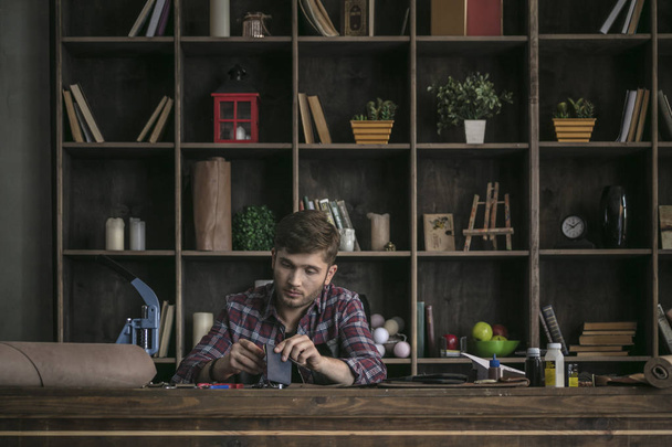 Young man leather manufacturer sitting behind wooden table and sewing wallet - Valokuva, kuva