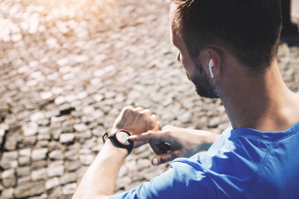 Γενειοφόρος νέος αθλητής με ακουστικά bluetooth αναλύει αποτέλεσμα της κατάρτισης για το smartwatch. Έννοια της σύγχρονης αθλητής με έξυπνο ρολόι και gadgets σε αστικό περιβάλλον - Φωτογραφία, εικόνα