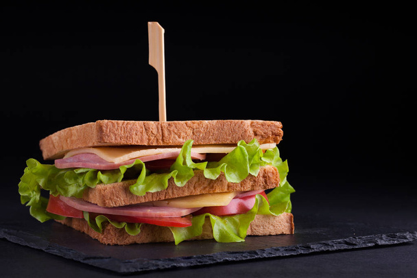 Sándwich grande de primer plano con jamón, queso, tomates y ensalada sobre pan tostado sobre un fondo oscuro
 - Foto, imagen
