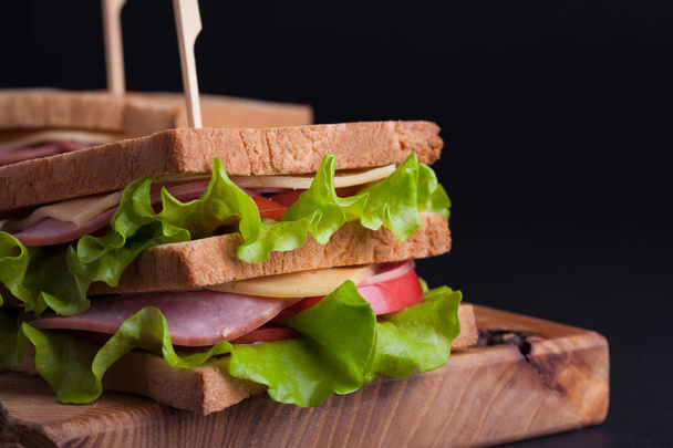 Lähikuva iso voileipä kinkkua, juustoa, tomaatteja ja salaattia paahdettua leipää tummalla pohjalla
 - Valokuva, kuva