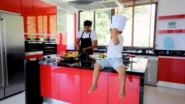 Тайська людина - шеф-кухар - приготування їжі, його маленький син в капелюсі шеф-кухаря неподалік сидять на столі в сучасному стилі домашньої кухні. Дитина їсть, тато, приготування їжі - Кадри, відео