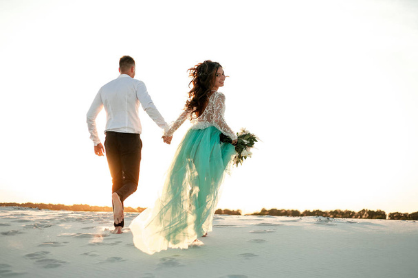 Νύφη και ο γαμπρός που τρέχουν στην άμμο στην έρημο και νύφη κοιτάζει πίσω. Πίσω - Φωτογραφία, εικόνα