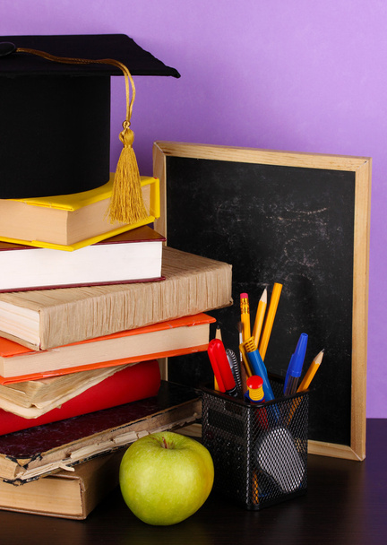 Books and magister cap against school board on wooden table on purple background - Foto, Imagen