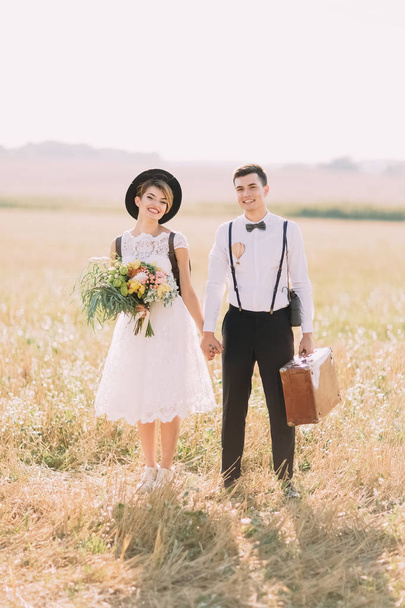 The front full-length portrait of the vintage dressed newlywed couple holding hands and smiling at the camera at the background of the sunny field. - Valokuva, kuva