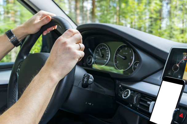 Male hands holding car steering wheel. Hands on steering wheel of a car driving near the lake. Man driving a car inside cabin. Smartphone in holder with  isolated white empty blank screen. Copy space - Photo, Image