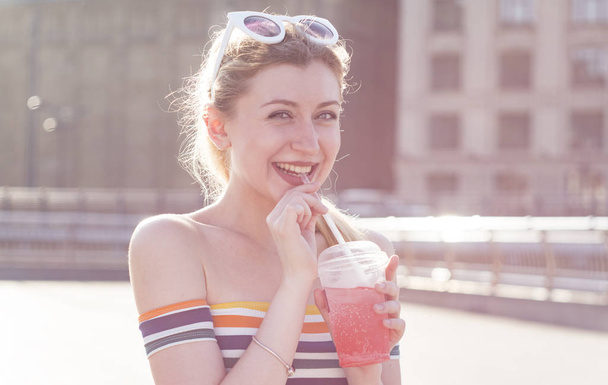kaunis nuori hymy blondi tyttö kaupungin kadulla aurinkoisena päivänä juo virkistävän hedelmäcocktailin jäällä
 - Valokuva, kuva